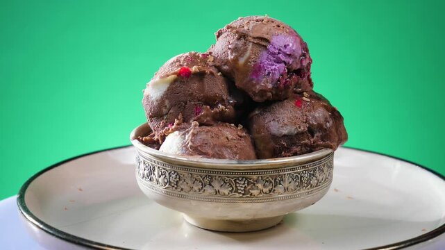 Balls of chocolate ice cream in a glass bowl, sprinkled with sweets, slowly rotate against a brightly colored background.