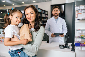 Happy woman holding little girl discussing medicine with friendly pharmacist in pharmacy