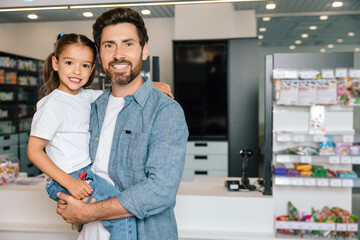 Naklejka premium Bearded father in casual denim shirt holding daughter smiling together in modern pharmacy