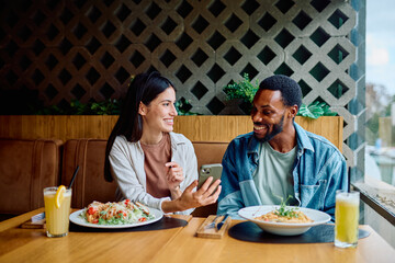 Diverse couple sitting at a restaurant table, smiling, and laughing while looking at a smartphone screen during their lunch date