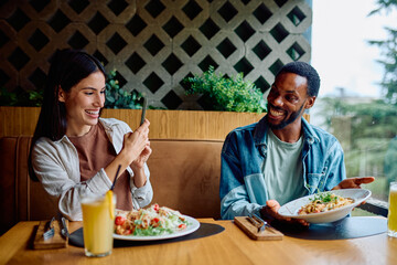 Smiling diverse couple enjoying dinner at a restaurant, woman taking a picture of the food with a smartphone while man holds the plate