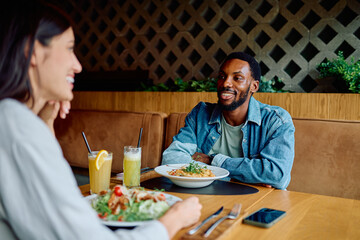 Young multicultural couple laughing over a casual daytime dinner at a modern restaurant, sharing pasta, salad and drinks, relaxed togetherness