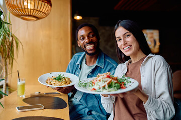 Smiling diverse couple enjoying lunch or dinner date at a restaurant, holding plates of pasta and salad