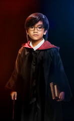 Naklejka premium A young boy dressed as a wizard holding a wand and old books, with dramatic red and blue lighting, resembling a fantasy character.