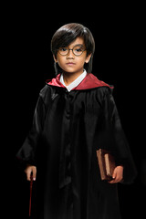 Fototapeta premium A young boy dressed as a wizard wearing black and red robe, holding a magic wand and old books while standing seriously against a black studio background. Fantasy, Halloween, or cosplay themes.