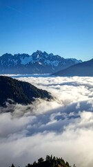 High-altitude view of a mountain valley shrouded in clouds.  Vast expanse of white clouds, dark shadows and hints of green below, with snow-capped peaks piercing the blue sky above