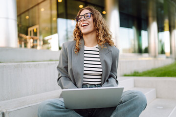 Portrait of a stylish woman with a laptop sitting on the steps in front of a modern office building. A beautiful woman in glasses works remotely outdoors. Freelancing concept, weekend.