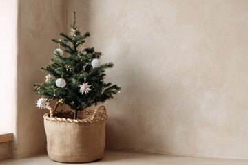 A small Christmas tree decorated with white ornaments and string lights stands in a rustic linen basket placed in a bright corner against a beige textured wall