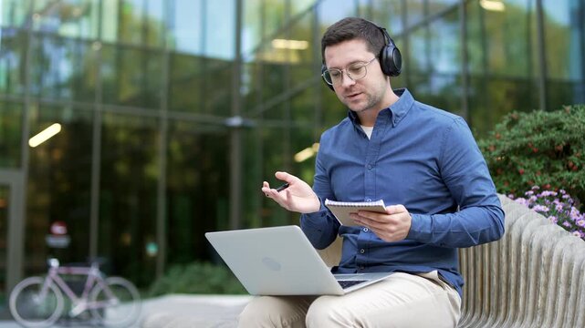 Confident businessman in headphones watching video call online conference notes in notebook sitting outside on a bench near office building. Worker communicates remotely at business meeting, training
