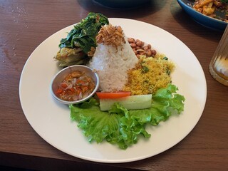 Balinese dish served on white plate featuring Bebek Betutu roast duck, accompanied by amound of rice topped with fried shallots. includes sauteed greens contain grated coconut, roasted peanuts, salad.