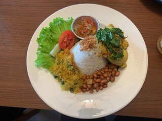 Balinese dish served on white plate featuring Bebek Betutu roast duck, accompanied by amound of rice topped with fried shallots. includes sauteed greens contain grated coconut, roasted peanuts, salad.
