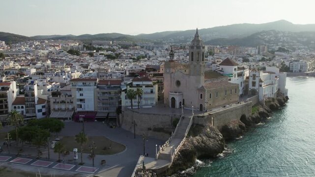 Aerial view of the Church of Sant Bartomeu i Santa Tecla, a baroque-style church with a bell tower, overlooking the blue waters, Sitges, Barcelona, Spain.