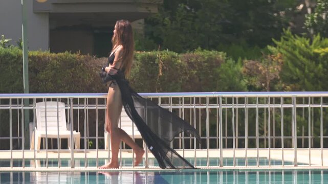 A beautiful woman, draped in a stylish black pareo, walks along the edge of a glistening pool on a sunny day. Her elegant stride and the clear, sunlit water create a perfect blend of summer luxury.