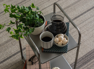 A glass of coffee, a pot of coffee, meringue in a bowl, a book, a pet flower on a metal table in the living room. Coffee break