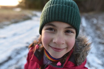 Smiling child in winter clothes outdoors. Close-up portrait of a cheerful girl in a green knitted hat and red winter jacket smiling outdoors on a cold day. Warm sunset light, snow on the ground, soft 