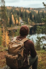 Stock photo from behind of a young male hiker with a backpack and beanie, seated on a rock and looking out at a stunning forest and lake with autumn foliage.