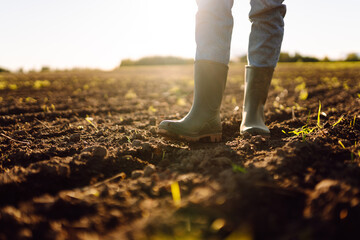 A close-up of a farmer's feet in green rubber boots in a field. A young female agronomist stands in...