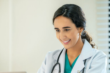 Woman female doctor portrait in white coat gown with stethoscope sit and smile to front. Medical student attractive friendly wear white coat smiling and work on table in patient room
