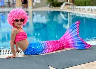 A cute little girl in a mermaid costume sits near the pool on a bright sunny day