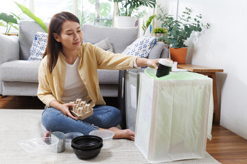 Young Asian woman sorting recyclable waste at home, putting plastic containers and paper packaging...