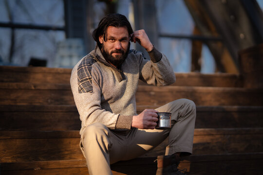 Man sitting on wooden steps wearing a sweater and holding a mug in a natural setting