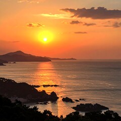 Sunrise over a tranquil coastline.  Sunrise paints the ocean and clouds in warm orange and yellow hues.  Silhouette of coastal hills and rocky outcrops