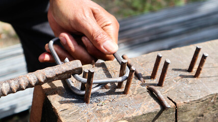 Worker manually bends steel rebar into stirrups using hammer and nail jig on wooden block. Ideal for construction, fabrication, and civil engineering process themes.
