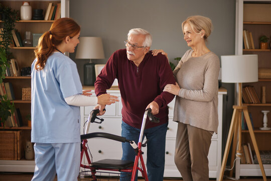 A young female nurse in scrubs helps an older man use his walker while his wife assists at their home. They are standing in a living room with shelves and a lamp.