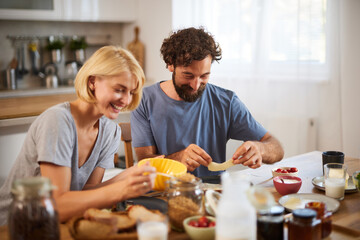 A couple smiles and engages at their kitchen table, preparing breakfast. The atmosphere is warm, with sunlight streaming in and a spread of food in front of them.