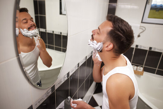 A man stands in a modern bathroom, looking into the mirror while applying shaving cream to his face. The tiles are black and white, adding a sleek look to the space.