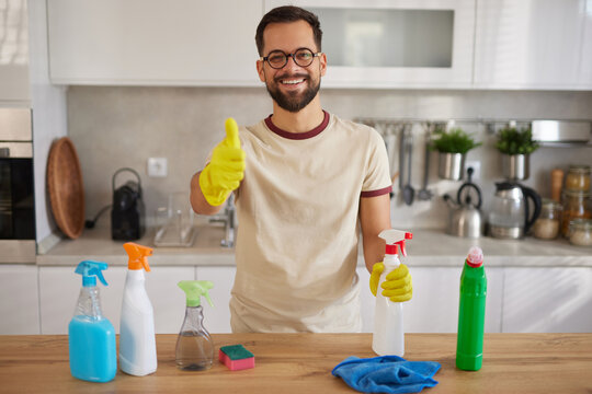 A cheerful man wearing yellow gloves stands in a modern kitchen. Surrounded by various cleaning products, he smiles and gives a thumbs up, showing satisfaction in his cleaning activity.