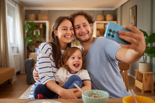 Smiling parents and their young child take a fun selfie while enjoying breakfast together in a warm and inviting living room filled with natural light.