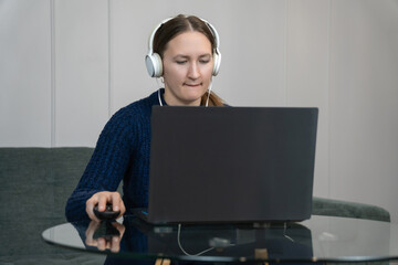 Young woman wearing headphones and working from home, typing on a laptop while sitting on a sofa, concentrating on her tasks in a comfortable remote work environment