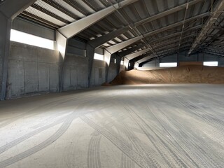Empty industrial warehouse with concrete walls and floor, bulk grain pile under metal roof with skylights