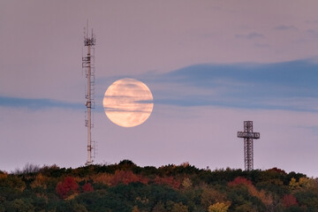 A supermoon rises above autumn treetops and a cross, with a radio tower silhouette. g.