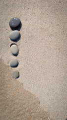 Vertical photo of smooth pebbles arranged in a vertical line on wet beach sand as a gentle sea wave touches them. Minimalist and peaceful seaside composition symbolizing balance harmony mindfulness