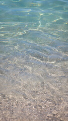 Vertical photo of transparent turquoise sea water with sunlight reflections and visible sandy seabed. The calm shallow water reveals natural textures and gentle ripples creating a peaceful summer