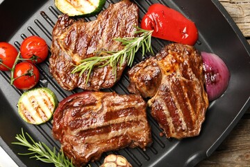 Pan with grilled beef steaks, rosemary and vegetables on wooden table, top view