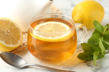 Aromatic tea in cup with lemons, mint, spoon and teapot on white marble table, closeup