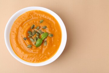 Delicious pumpkin soup with seeds and basil in bowl on beige table, top view. Space for text