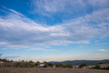 A rural Bulgarian landscape unfolds beneath a dramatic sky, with red-roofed houses nestled among dry fields and scattered trees, framed by rolling hills and textured clouds.