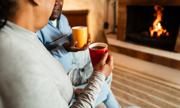 African senior couple drinking hot chocolate in front of cozy fireplace at home - Winter lifestyle and holidays concept - Focus on woman's hand - Powered by Adobe