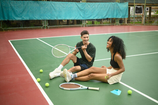 Two diverse athletes resting together on a tennis court after a training session, showing teamwork and friendship