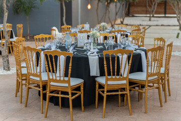 A beautifully arranged dining area features round tables with black tablecloths and gold chairs....