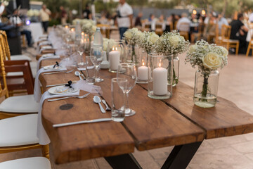 Decorated wooden tables are set with glasses, cutlery, and floral arrangements, creating a warm atmosphere for a gathering at an outdoor venue during sunset.