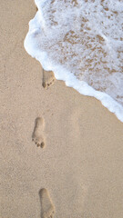 Vertical close-up of footprints on wet yellow sand as a gentle wave approaches, highlighting sand texture and white foam. Ideal for beach, summer, tropical, and relaxation concepts.