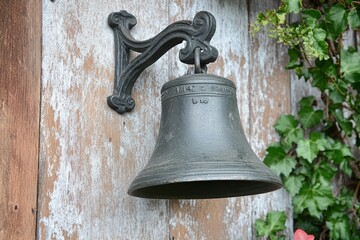 Obraz premium Old cast iron bell hanging on weathered wooden wall with green ivy climbing around. Rustic outdoor decoration for house entrance.