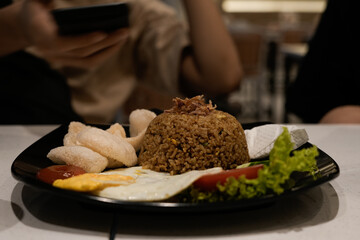  hearty Indonesian fried rice dish served with a sunny-side egg, crispy prawn crackers, fresh vegetables, and tomato slices on a sleek black plate