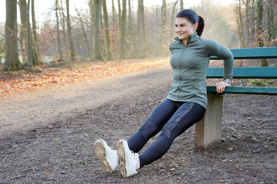 Athletic young woman doing stretching exercises at a park bench on a sunny winter morning in a forest