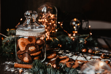 A rustic Christmas holiday setting featuring a jar of homemade cookies, surrounded by cinnamon sticks and festive lights. Pine branches add a cozy, warm atmosphere perfect for the winter season.